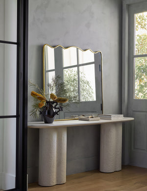 The rook mantel mirror sits atop a white sideboard with pedestal legs next to a window