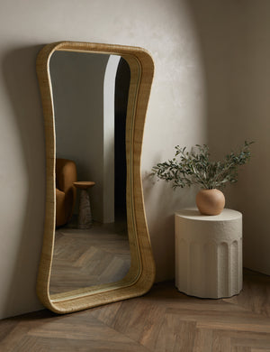 Howell wicker frame floor mirror leaning against a wall next to a round side table with a decorative vase.