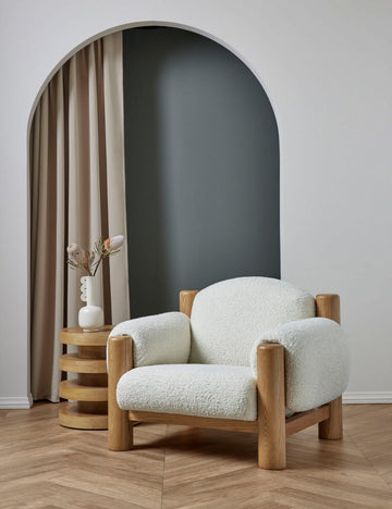 | The James faux sherpa accent chair sits in a studio next to a four-tiered natural side table with a white vase
