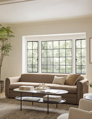 The Eleanor toffee brown sofa sits in front of a big window behind an oval coffee table and atop a distressed rug
