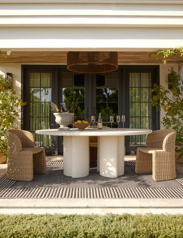 #color::natural | Rodrigo sculptural oval outdoor dining table styled with two wicker outdoor dining chairs in a covered outdoor space.