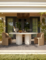 #color::natural | Rodrigo sculptural oval outdoor dining table styled with two wicker outdoor dining chairs in a covered outdoor space.
