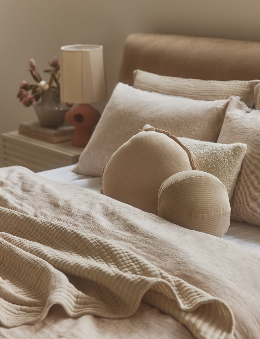 #color::light-natural | Arlo linen flange trim round pillow styled with other pillows on a bed.