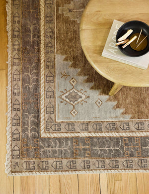 Bird’s-eye view of the bottom left corner of the Kehoe desert palette geometric floor rug sitting on a natural hardwood floor with a natural wood coffee table with a book and centerpiece bowl atop it.