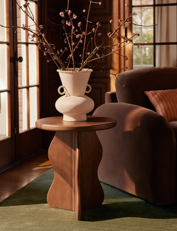 #color::dark-brown | The Telle wavy leg solid wood round side table by Aimee Song with a ceramic vase on top styled next to an accent chair