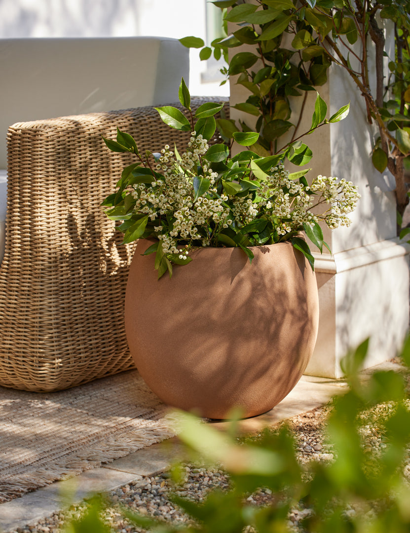 #size::medium | Kenna medium rounded fiberstone planter with plant outside beside a wicker sofa.