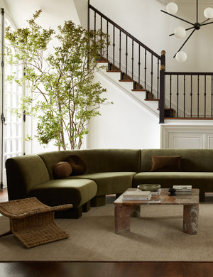The Heritage moss gray rug lays in a living room under a olive green velvet sectional, a woven ottoman, and a stone coffee table