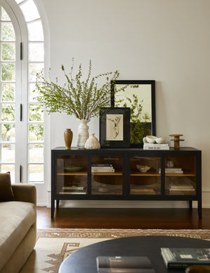 The Morey glass front black curio sideboard cabinet in a living room with various vases and artwork styled on top