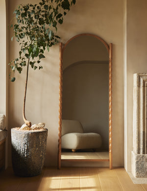 Topia arched carved wood frame floor mirror by Ginny Macdonald in natural leaned against a wall next to a decorative plant.