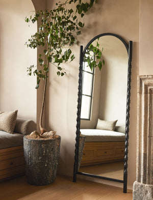 Topia arched carved wood frame floor mirror by Ginny Macdonald in black leaning against a wall next to a decorative plant.