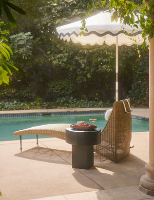Armas black monolithic round outdoor side table by Sarah Sherman Samuel sitting next to a wicker chaise by the poolside.