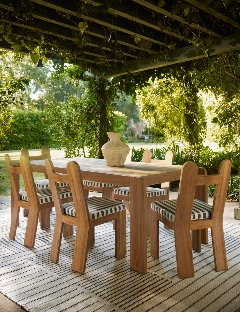 #color::natural-teak | Six Abbot solid teak sculptural outdoor dining chair by Sarah Sherman Samuel surround a teak outdoor dining table under a pergola.