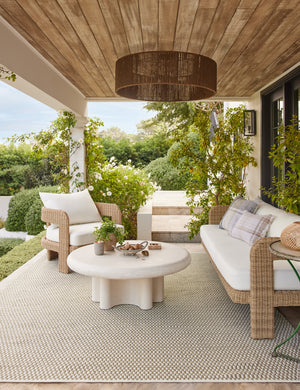 Ruiz round cement outdoor coffee table with a wicker outdoor sofa and chair in a covered outdoor space.