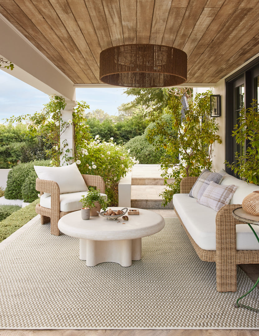 #color::ivory | Ruiz round cement outdoor coffee table with a wicker outdoor sofa and chair in a covered outdoor space.