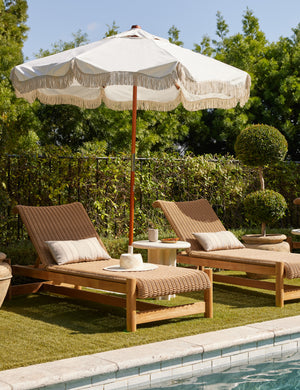 Ruiz round cement outdoor side table between two wicker chaise lounges and a white umbrella by the pool.