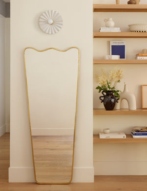 The Rook golden full length mirror hangs on a white wall above a ripple-shaped accent chair next to a shelf with decor