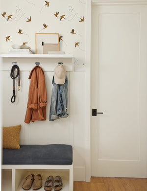 The Sparrow brown wallpaper by Rylee and Cru is in an entryway aboved an accented wall with a white coat rack and a blue cushioned bench.