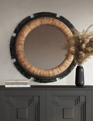 Eloy round woven natural fiber wall mirror hung above a black console table.