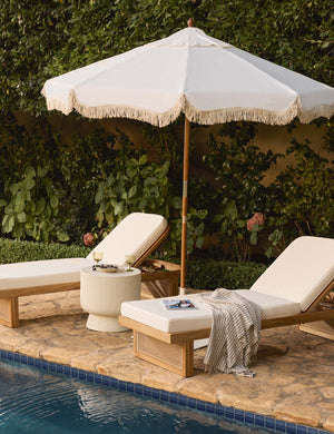 The Market vintage white umbrella by business and pleasure co with a cotton fringe sits beside a pool between two white lounge chairs