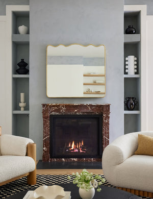 The rook mantel mirror hangs above a burgundy marble fireplace in between two boucle chairs and wall shelves