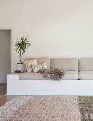 The Terra handcrafted textured multicolored floor rug by Élan Byrd sits in a living room with a gray linen cushioned bench, a gray sheepskin throw, and green and terracotta throw pillows