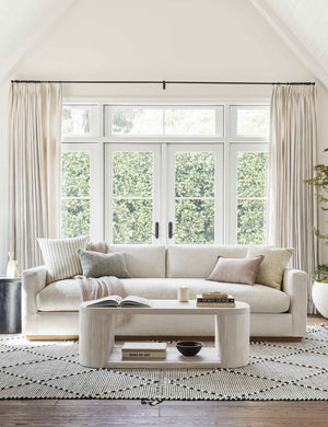 The Luna white-washed oak oval coffee table sits in a well-lit living room atop a white and black patterned rug in front of a white couch with long windows and french doors in the background.