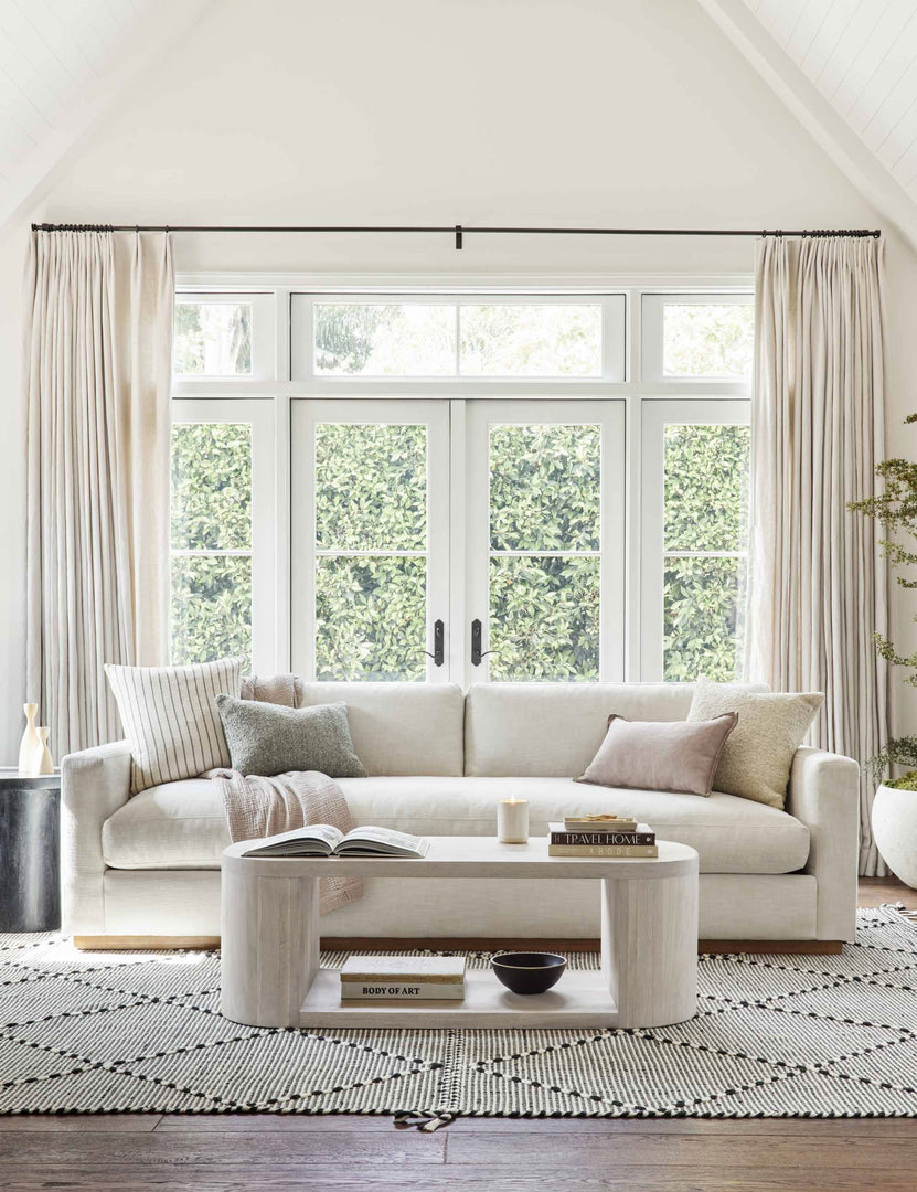 #color::washed-oak | The Luna white-washed oak oval coffee table sits in a well-lit living room atop a white and black patterned rug in front of a white couch with long windows and french doors in the background.