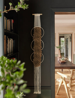  The Studio Nom Lwazi light brown woven Wall Hanging by nom hangs in a black painted room next to a bookshelf and a dining room in the background