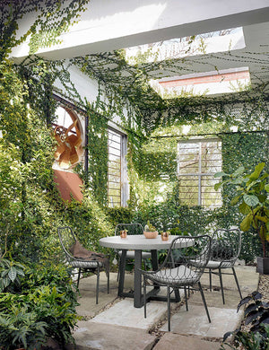 The Agatha indoor and outdoor dining table with lavastone top and black iron base sits in a covered outdoor space with ivy-covered walls and black woven dining chairs