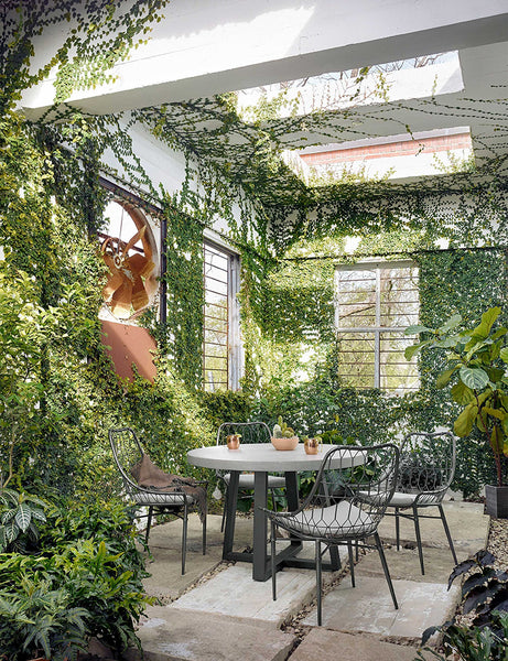 | The Agatha indoor and outdoor dining table with lavastone top and black iron base sits in a covered outdoor space with ivy-covered walls and black woven dining chairs 