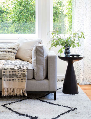 The Bridget bronze pedestal side table with mirror ash glass top sits in a living room with a gray linen sofa, a plush patterned rug, and a geometric throw blanket