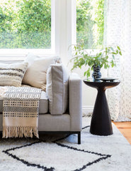 | The Bridget bronze pedestal side table with mirror ash glass top sits in a living room with a gray linen sofa, a plush patterned rug, and a geometric throw blanket