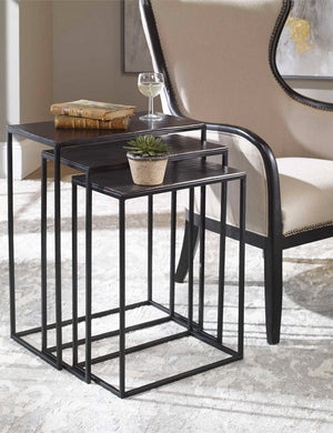 The Loletta black nesting tables are partially nested under each other, sitting in a living room next to a black wooden framed accent chair atop a gray patterned rug