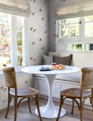 The Sparrow brown wallpaper by Rylee and Cru is in a dining nook with a white sculptural dining table, brown woven chairs that are surrounded by large colonial style windows.