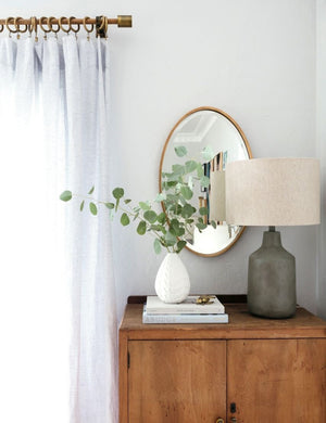 The Merci gold framed oval mirror is hung against a white wall behind a wooden side table and a white textured vase