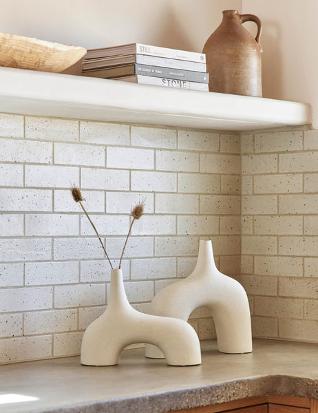 #color::white #size::small | The Leonor sculptural arched matte white ceramic Vase in its small and large size sitting atop a granite countertop in a kitchen with a subway tile backsplash, a stack of books, and a wooden centerpiece bowl
