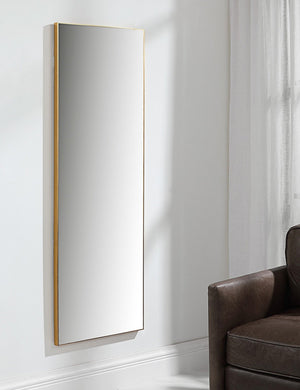 The Shea rectangular gold framed full length floor mirror hangs on a wall in a living room with a brown leather chair
