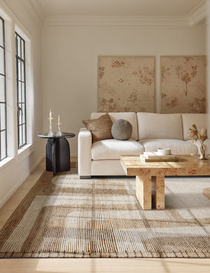 The Abode geometric two-toned kand-knotted floor rug by Élan Byrd with woven border sits in a living room with a burl wood coffee table, floral wall art, and a plush beige linen sofa