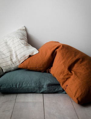 The set of two european flax linen cedar orange pillowcases by cultiver lay in a pile of pencil stripe and bluestone pillows