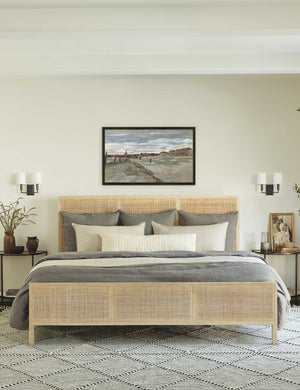 The Hannah bed with light wood cane bed frame sits beneath a landscape painting with two double sconces and a black-and-white patterned rug below it.