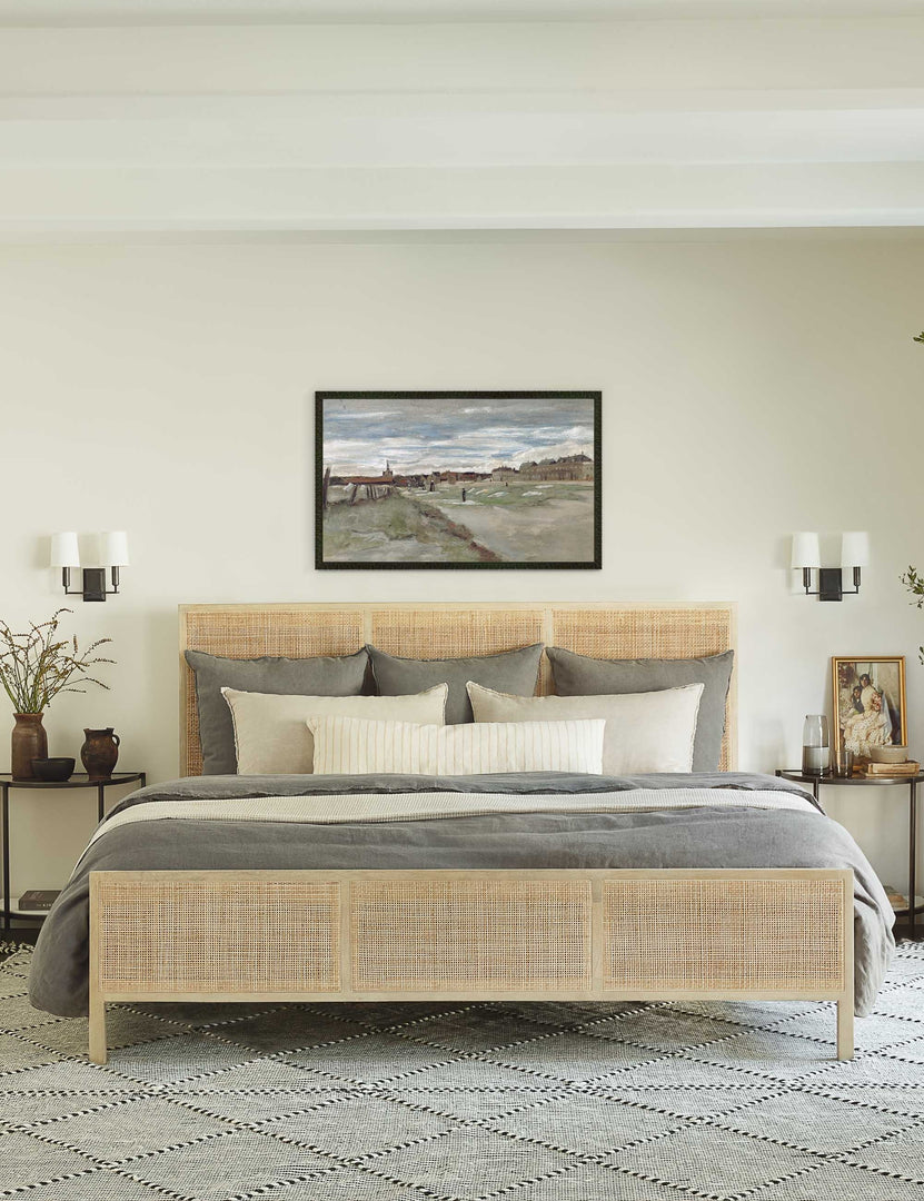 #size::king #size::queen #color::natural | The Hannah bed with light wood cane bed frame sits beneath a landscape painting with two double sconces and a black-and-white patterned rug below it.