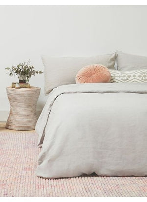 The Set of two european flax linen smoke gray pillowcases by cultiver sit on a bed with other smoke gray cultiver linens in a bedroom with a pink textured rug and a wooden ringed nightstand