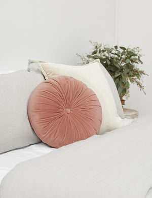 The Monroe coral pink velvet round pillow sits on a bed with gray and natural linens