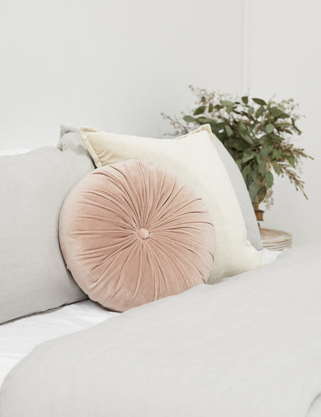 #color::rosewater | The Monroe rosewater pink velvet round pillow sits on a bed with gray and natural linens