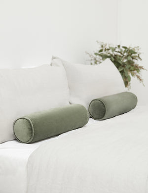 Two Sabine moss green velvet cylindrical bolster pillows sit on a bed with gray linens with a plant in the background