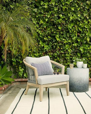 The Pira gray and white ombre indoor and outdoor side table sits in an outdoor space with a gray linen accent chair, a black striped rug, and an ivy wall
