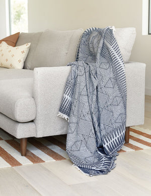 The Giana blue and white geometric throw blanket sits in a living room laying atop a gray linen sofa with a orange and white striped rug underneath
