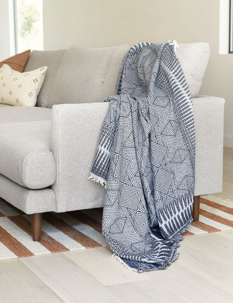 | The Giana blue and white geometric throw blanket sits in a living room laying atop a gray linen sofa with a orange and white striped rug underneath