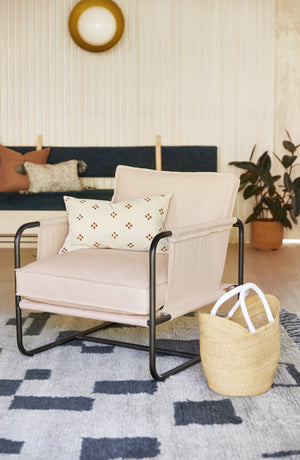 The Alena accent chair with cream linen cushions and black metal frame sits in a living room with a black and gray plush patterned rug, an orange and beige patterned throw pillow, and a woven cane basket