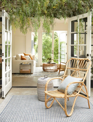 The Sonya gray and blue geometric indoor and outdoor rug lays in an outdoor space with a woven jute side table and a woven accent chair with a cream throw pillow